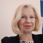 Smiling woman with blonde hair in a black blazer against a light background.