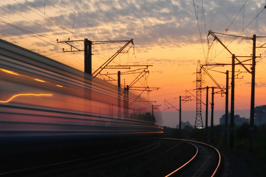 Train speeding down railway tracks at sunset, with silhouetted power lines against a colorful sky.